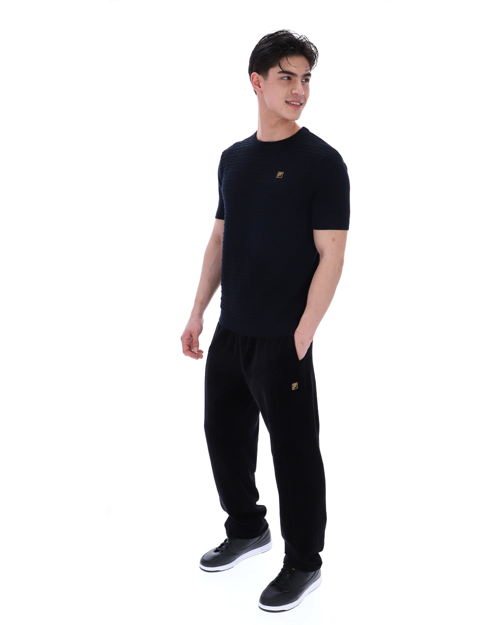 Man wearing a black t-shirt with a small FILA Gold logo on the chest with matching bottoms on a white background
