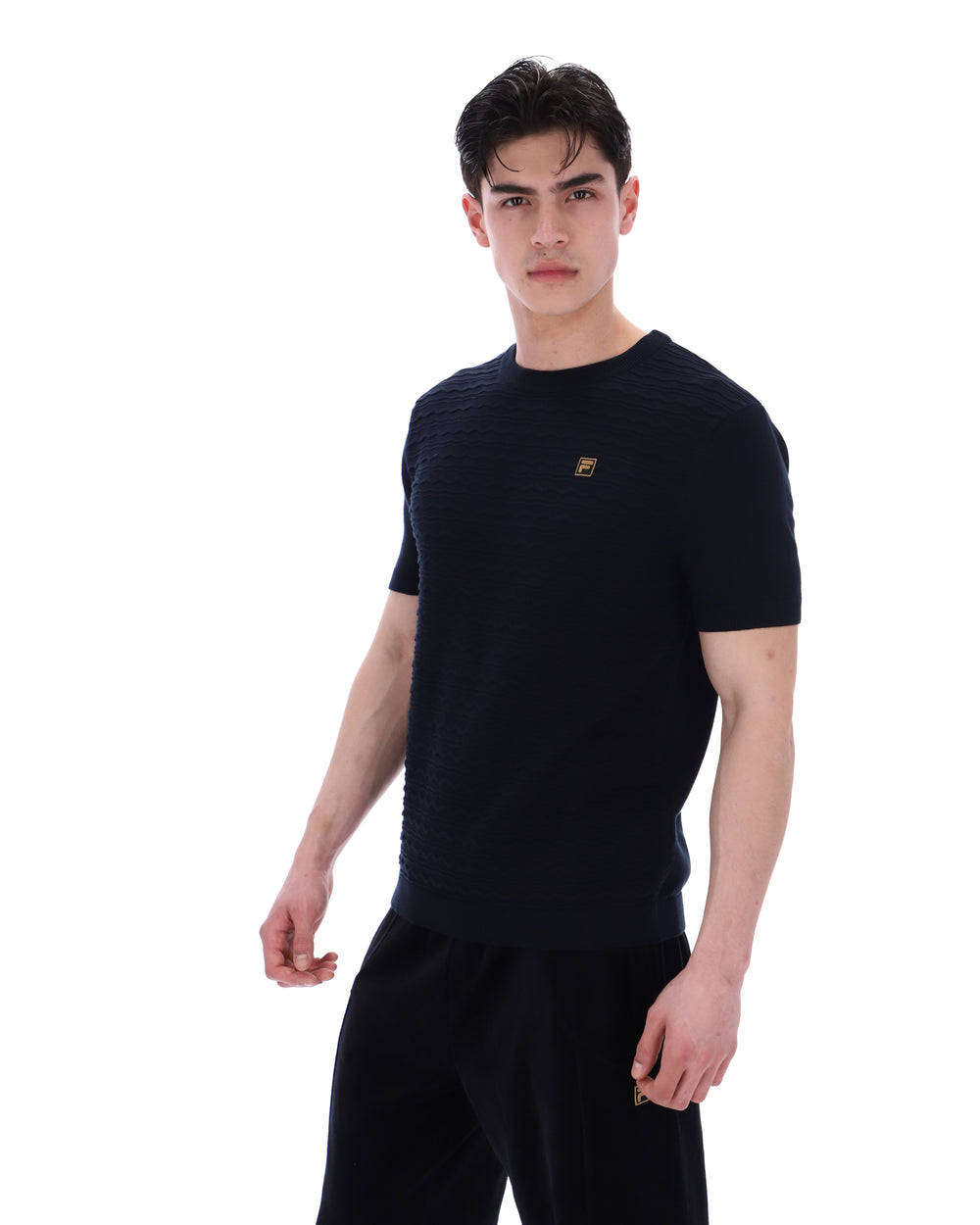 Man wearing a black t-shirt with a small FILA Gold logo on the chest with matching bottoms on a white background