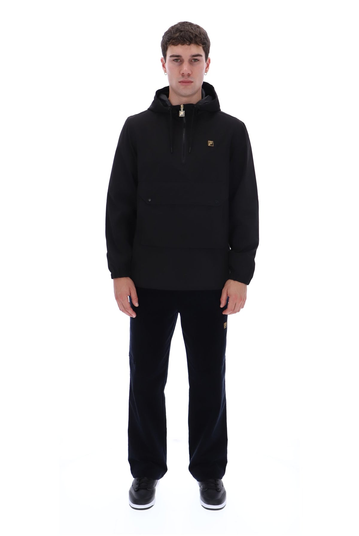 A man modeling a black hooded pullover jacket with a half-zip front and a small gold chest logo, paired with black pants and black sneakers, against a plain white background.