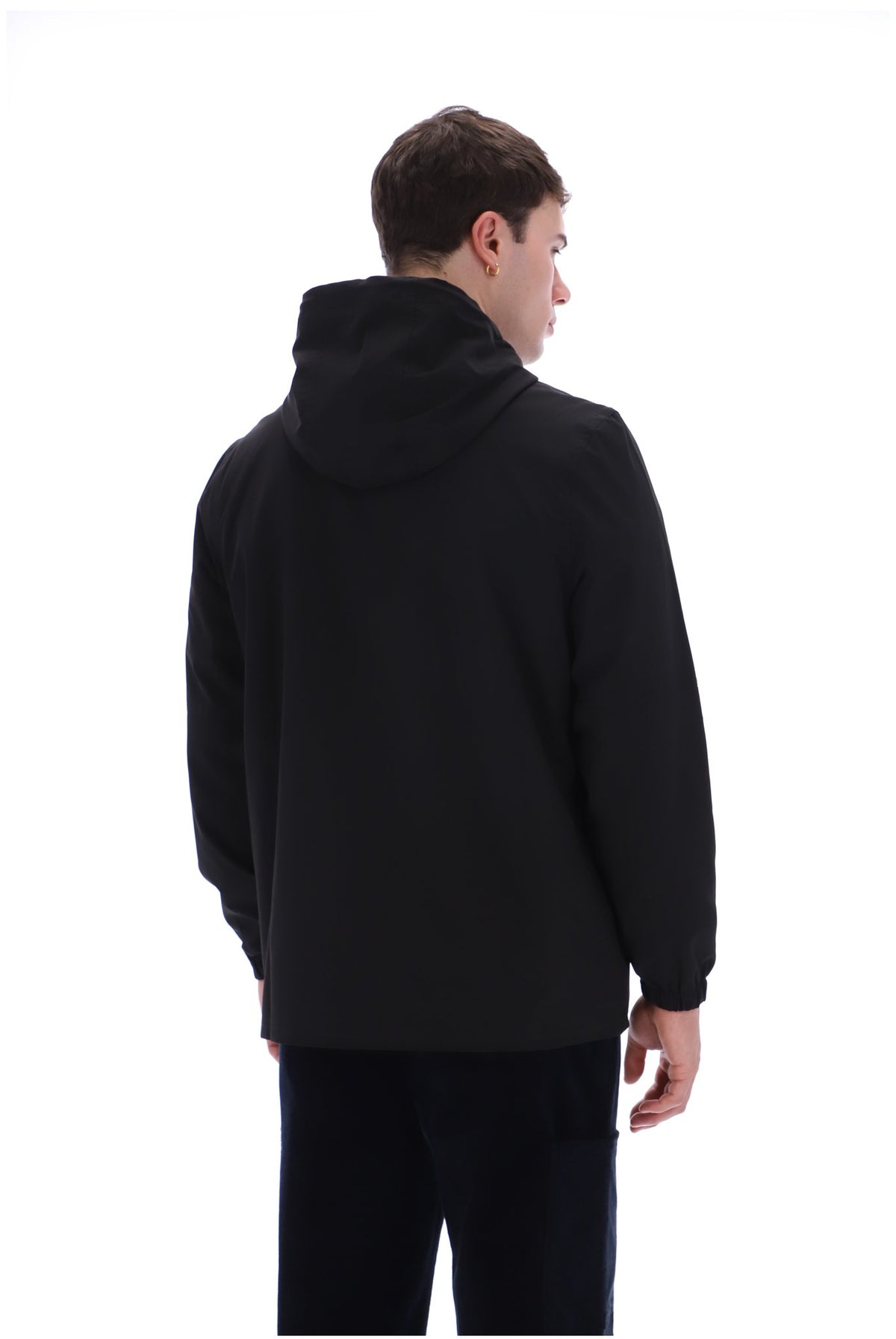 A man viewed from the back wearing a black hooded jacket and dark pants against a plain white studio background.