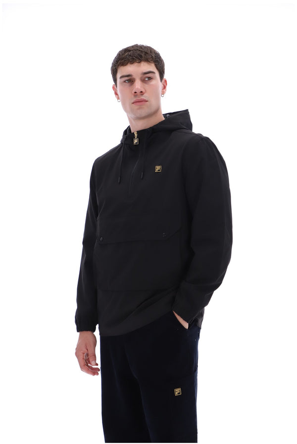 A man wearing a black pullover hoodie with a small gold logo on the chest, paired with black pants featuring a matching gold logo near the lower leg, set against a plain white background.
