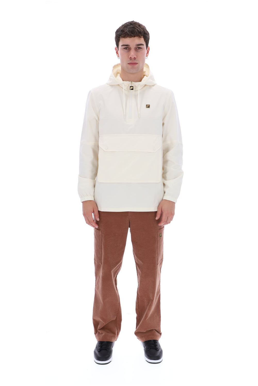 A man modeling a color-blocked hooded pullover jacket in cream and off-white, paired with brown pants and black sneakers, shot against a solid white background.