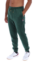 A person wearing dark green jogger sweatpants with a drawstring waist, side pocket, and a small Fila logo patch on the left thigh, paired with white sneakers against a plain white background.