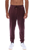 A man wearing dark plum corduroy jogger sweatpants with a drawstring waist and white sneakers, shown from the waist down against a plain white background.