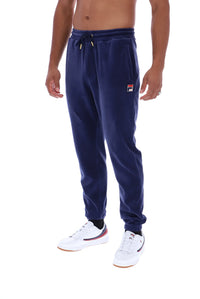A man wearing navy blue sweatpants with a small red, white, and blue Fila logo patch on the left thigh, paired with white sneakers, shown against a plain white background.
