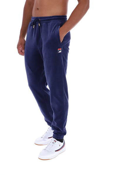 A person wearing navy blue jogger sweatpants with a drawstring waist and a small Fila logo patch on the left thigh, paired with white sneakers against a plain white background.