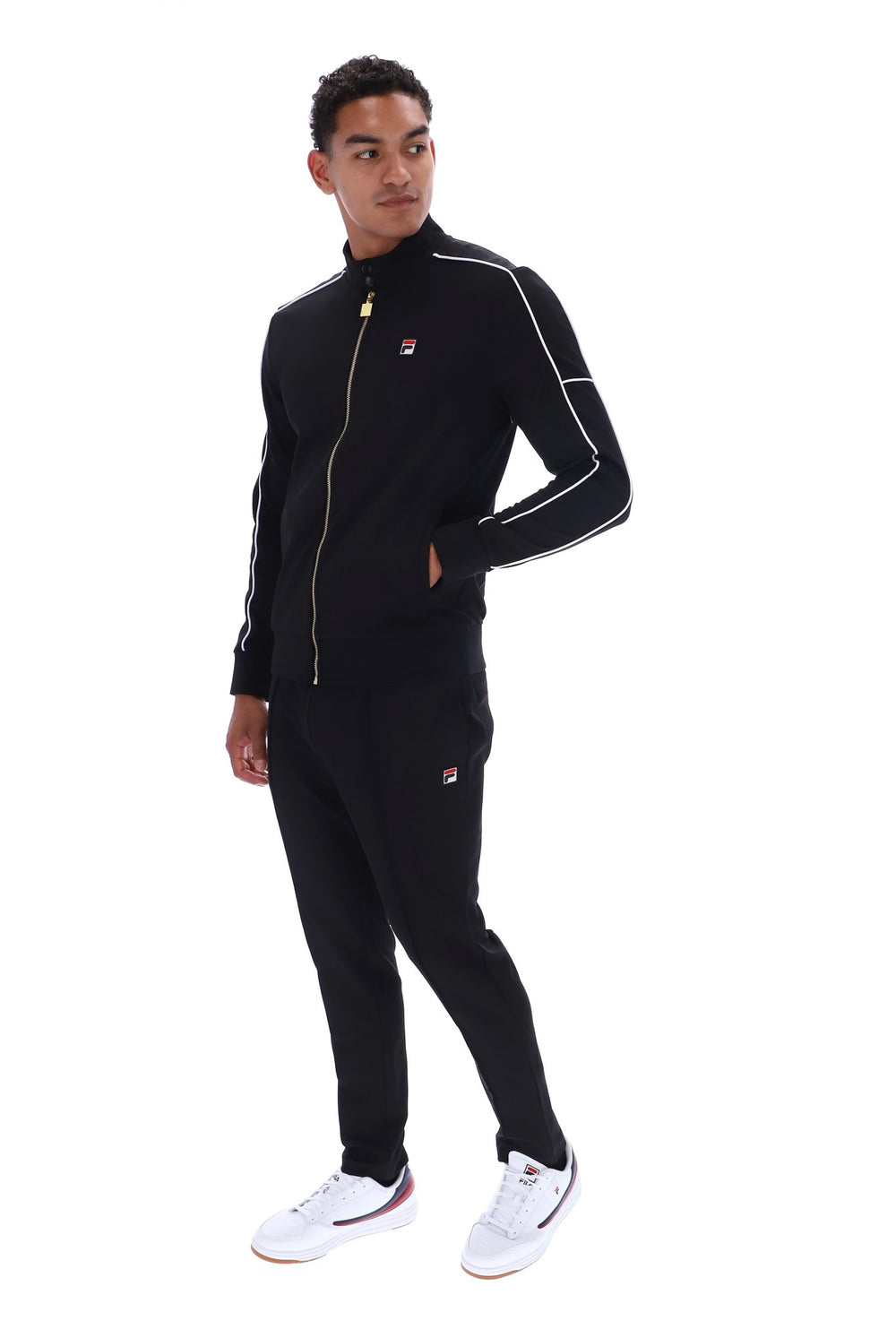 A man modeling a black Fila tracksuit with white piping and gold zipper accents, paired with white sneakers, against a plain white background.