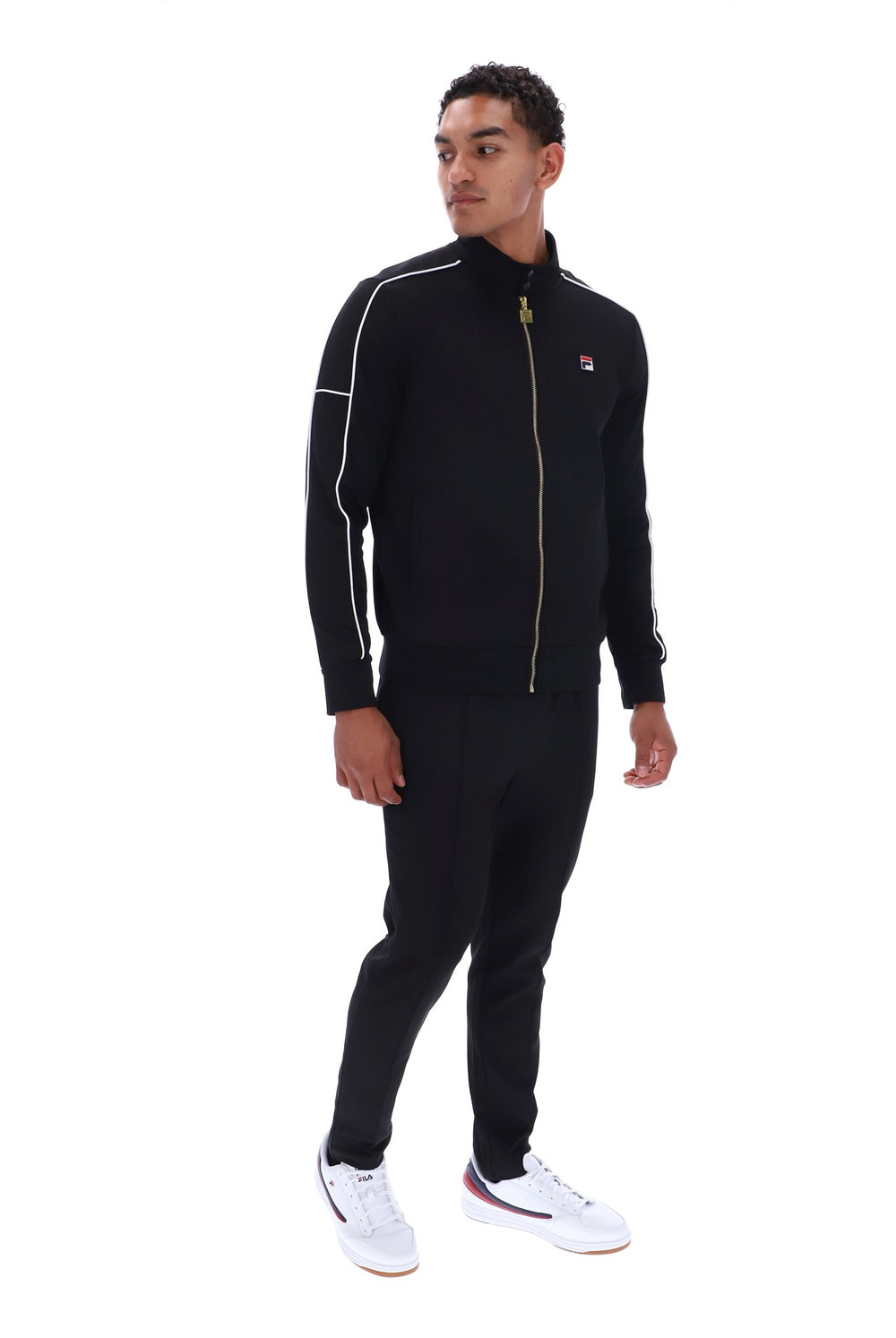 A man modeling a black Fila tracksuit, featuring a zip-up jacket with white piping and a small red, white, and blue logo on the chest, paired with matching black joggers and white sneakers, shot against a solid white background.