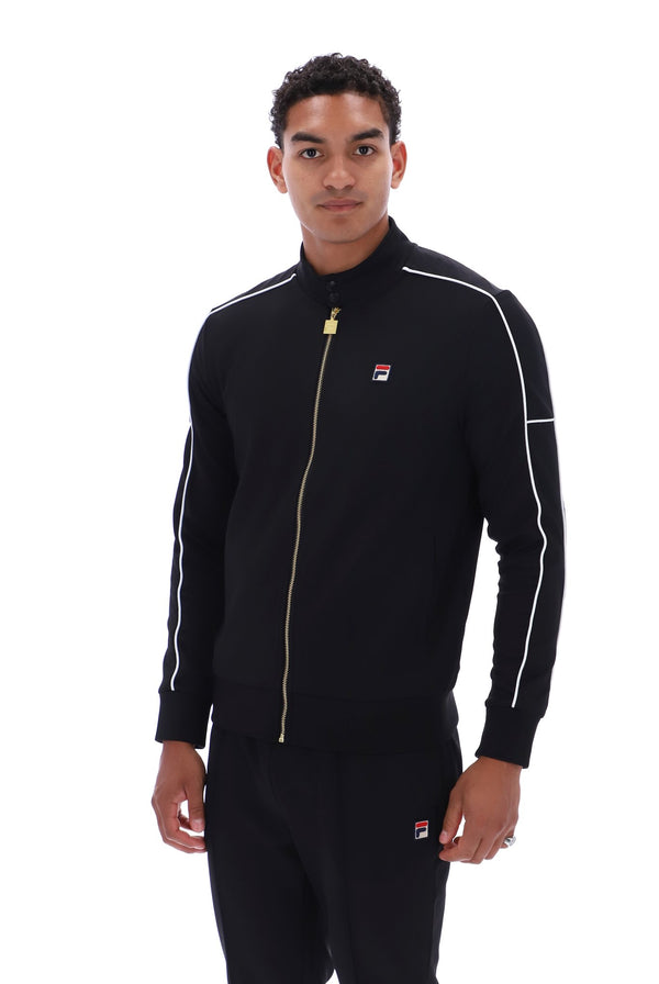 A man wearing a black Fila track jacket with white piping and a gold zipper, paired with matching black track pants, against a plain white background.
