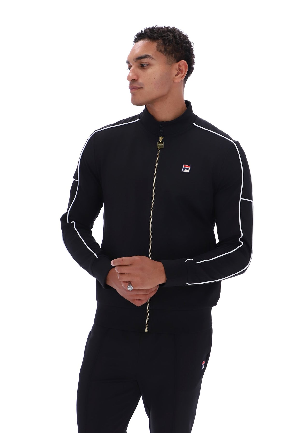 A man wearing a black Fila zip-up track jacket with white piping and a gold zipper, paired with matching black track pants, shot against a solid white background.