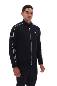 A man wearing a black Fila track jacket with white piping and a gold zipper, paired with black track pants, against a solid white background.