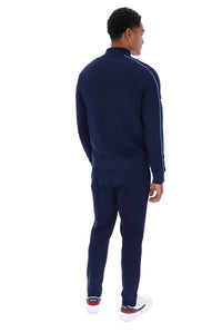 A man shown from the back wearing a navy blue tracksuit with white side stripes, navy pants, and white sneakers with red accents, set against a plain white background.