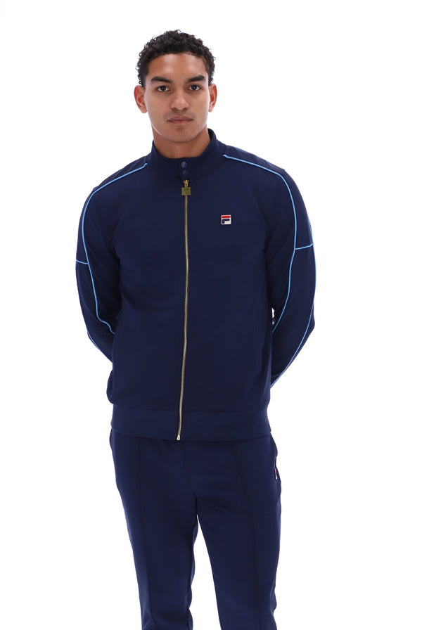 A man modeling a navy blue Fila tracksuit, featuring a zip-up jacket with light blue piping and a gold zipper, paired with matching navy pants, against a solid white background.