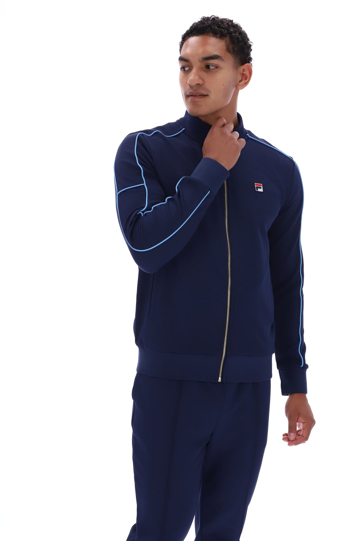 A man modeling a navy blue Fila tracksuit with light blue piping details and a gold zipper, set against a plain white background.