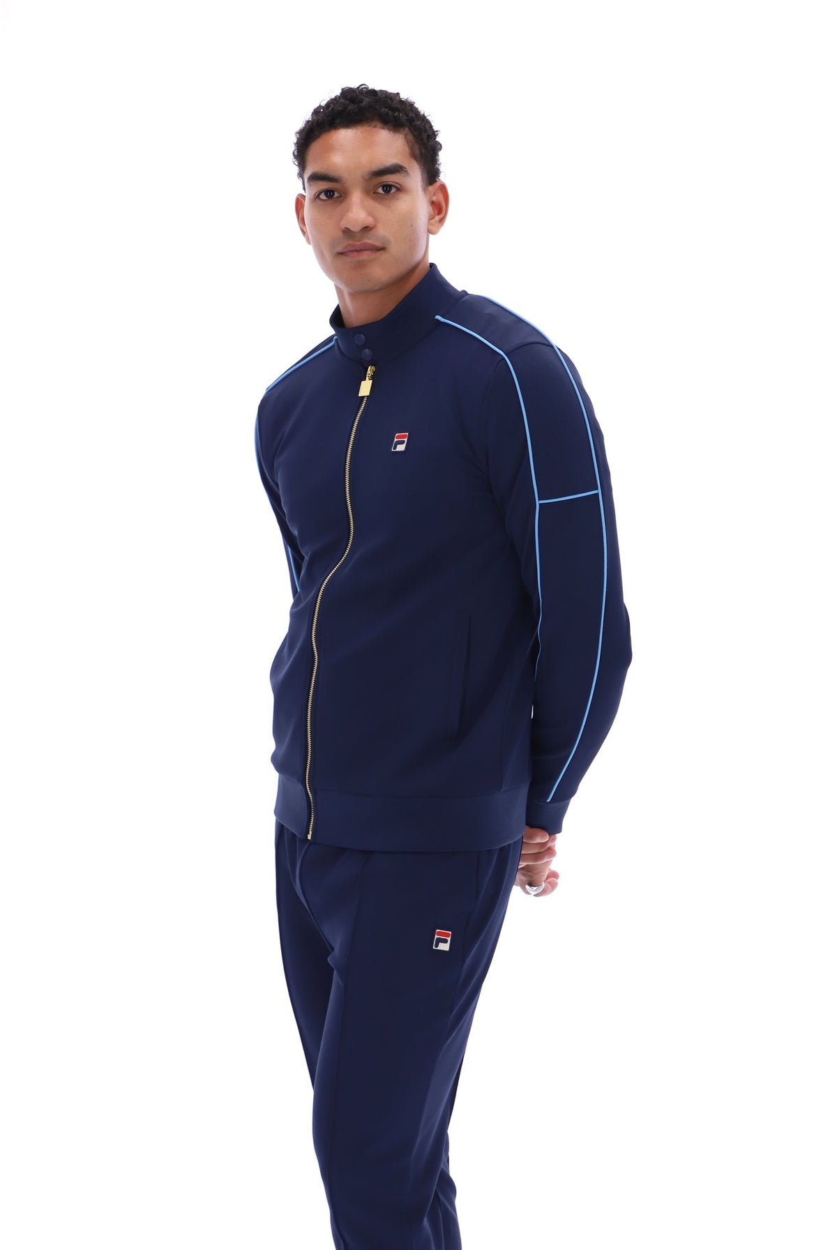 A man modeling a navy blue Fila tracksuit with light blue piping and a gold zipper, shot against a solid white background.