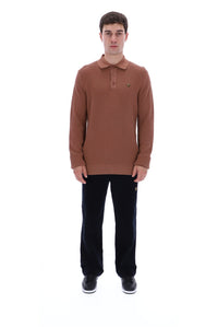 A man modeling a textured, long-sleeve polo shirt in a warm brown or rust color, paired with black trousers and black sneakers, against a solid white background.