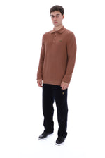 A man modeling a textured brown long-sleeve polo shirt with black trousers and black sneakers, standing against a plain white background.
