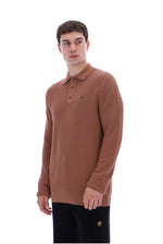 A man wearing a brown textured knit polo sweater with a small green embroidered logo on the chest, paired with black trousers, photographed against a solid white background.