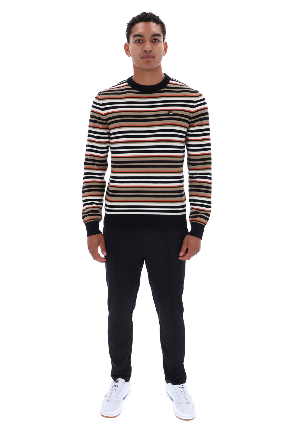 A man modeling a long-sleeve crewneck sweater with horizontal stripes in brown, black, and white, paired with black trousers and white sneakers, against a plain white background.