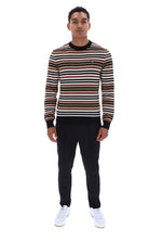A man modeling a long-sleeve crewneck sweater with horizontal stripes in brown, black, and white, paired with black trousers and white sneakers, against a plain white background.