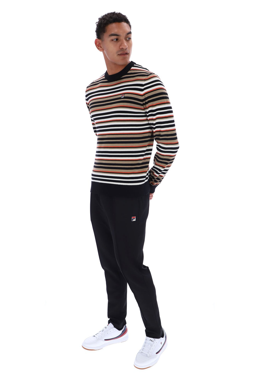 A man wearing a long-sleeve crewneck sweater with horizontal stripes in black, beige, and white, paired with black sweatpants and white sneakers, standing against a plain white background.