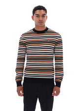 A man wearing a long-sleeve crewneck sweater with horizontal stripes in black, white, tan, and brown, paired with black trousers, photographed against a solid white background.