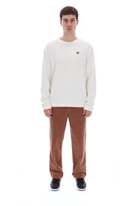 A man modeling a cream long-sleeve crewneck sweatshirt with a small brown logo on the left chest, paired with brown corduroy pants and black sneakers, against a solid white background.