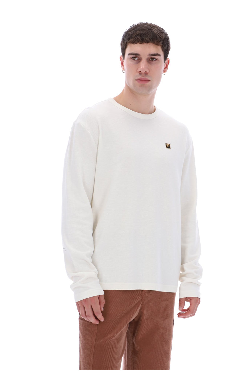 A man wearing a cream-colored long-sleeve crewneck sweatshirt with a small brown square logo on the left chest, paired with brown pants, photographed against a solid white background.