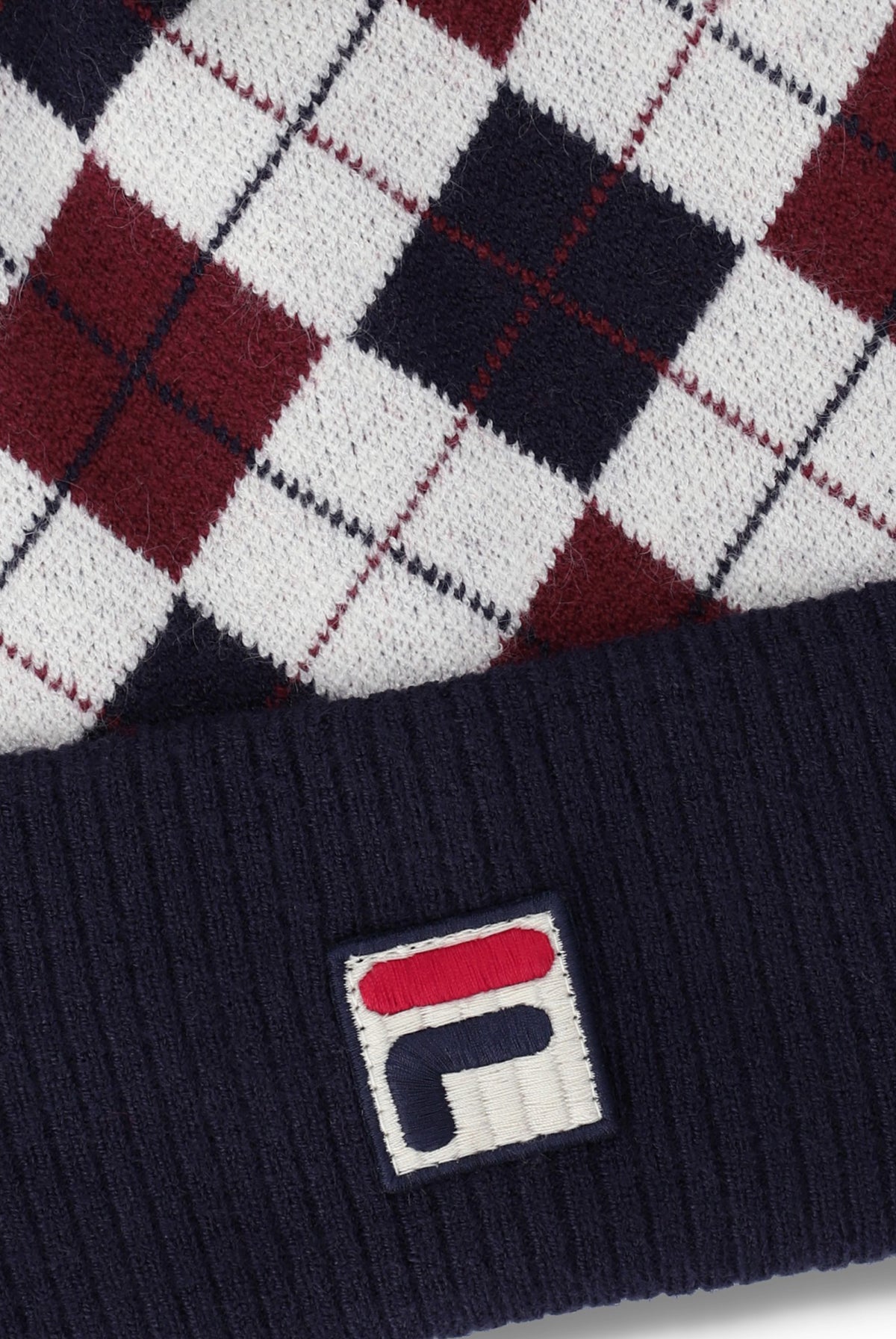 Close-up of a knit beanie with an argyle pattern in white, navy, and burgundy, featuring a navy ribbed cuff and a small embroidered Fila logo patch.