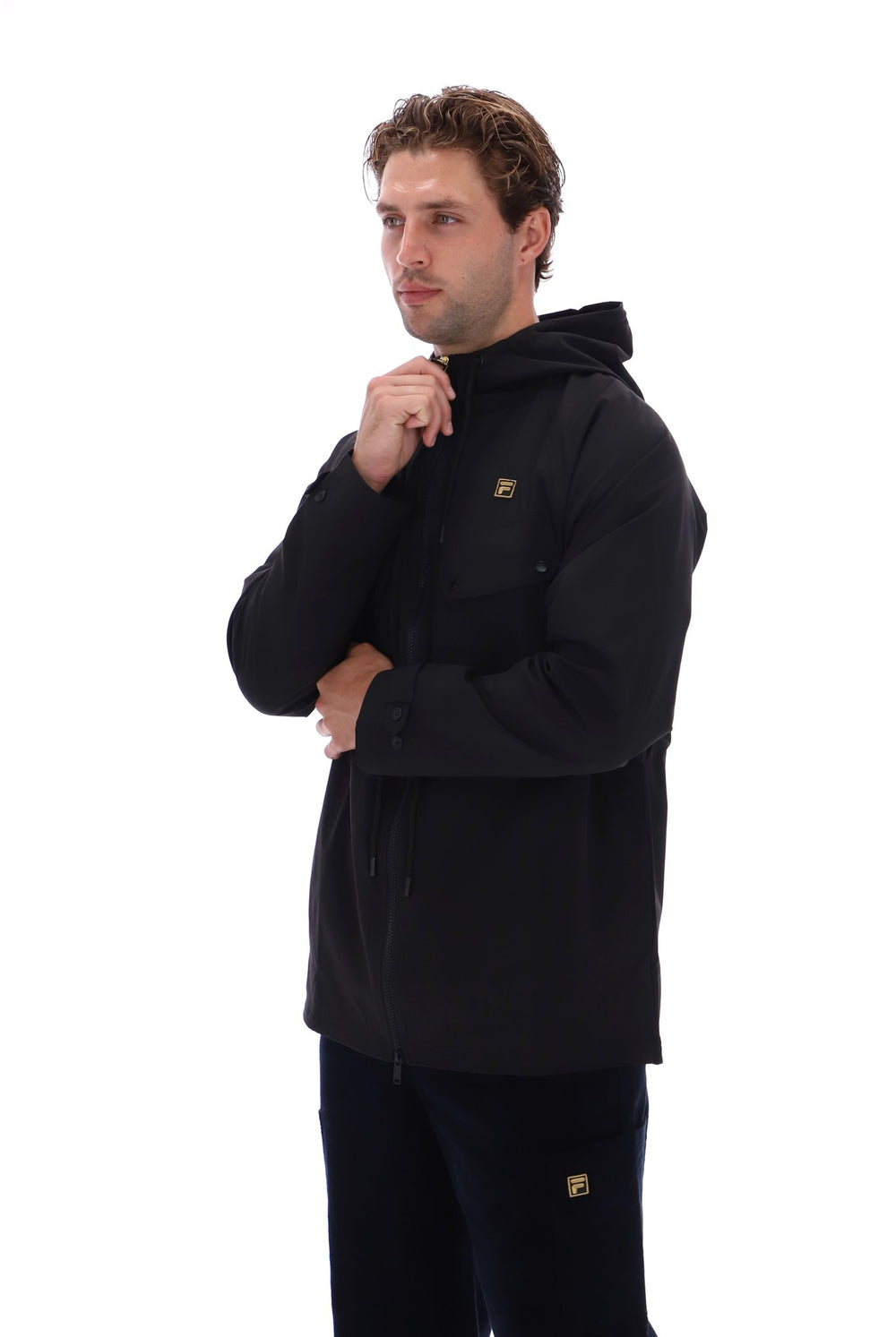 A man wearing a black hooded jacket with a small gold logo on the chest, paired with black pants, photographed against a solid white background.