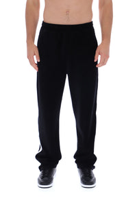 A man wearing black sweatpants with white side stripes and black sneakers, shown from the waist down against a plain white background.