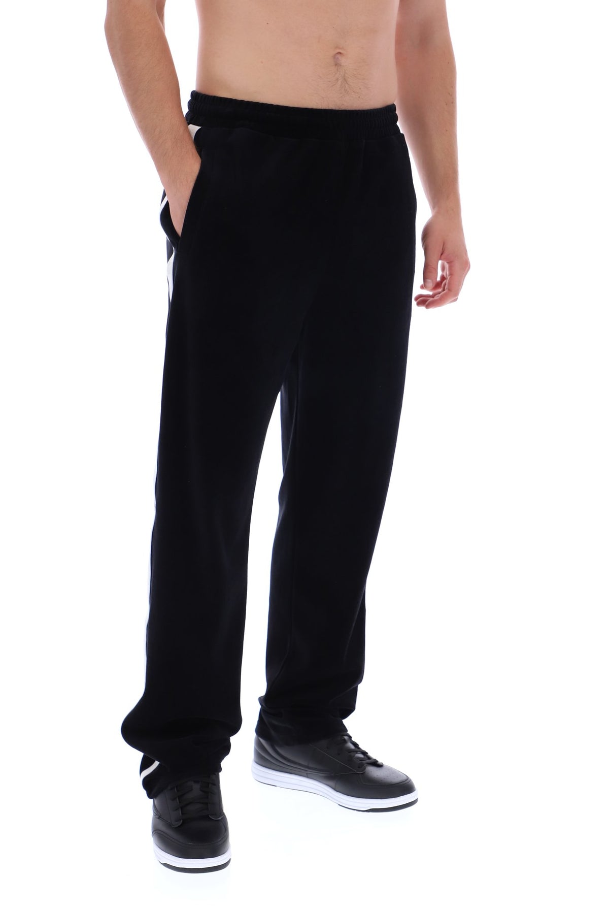 A man wearing black sweatpants with white side stripes and black sneakers, shown from the waist down against a white background.