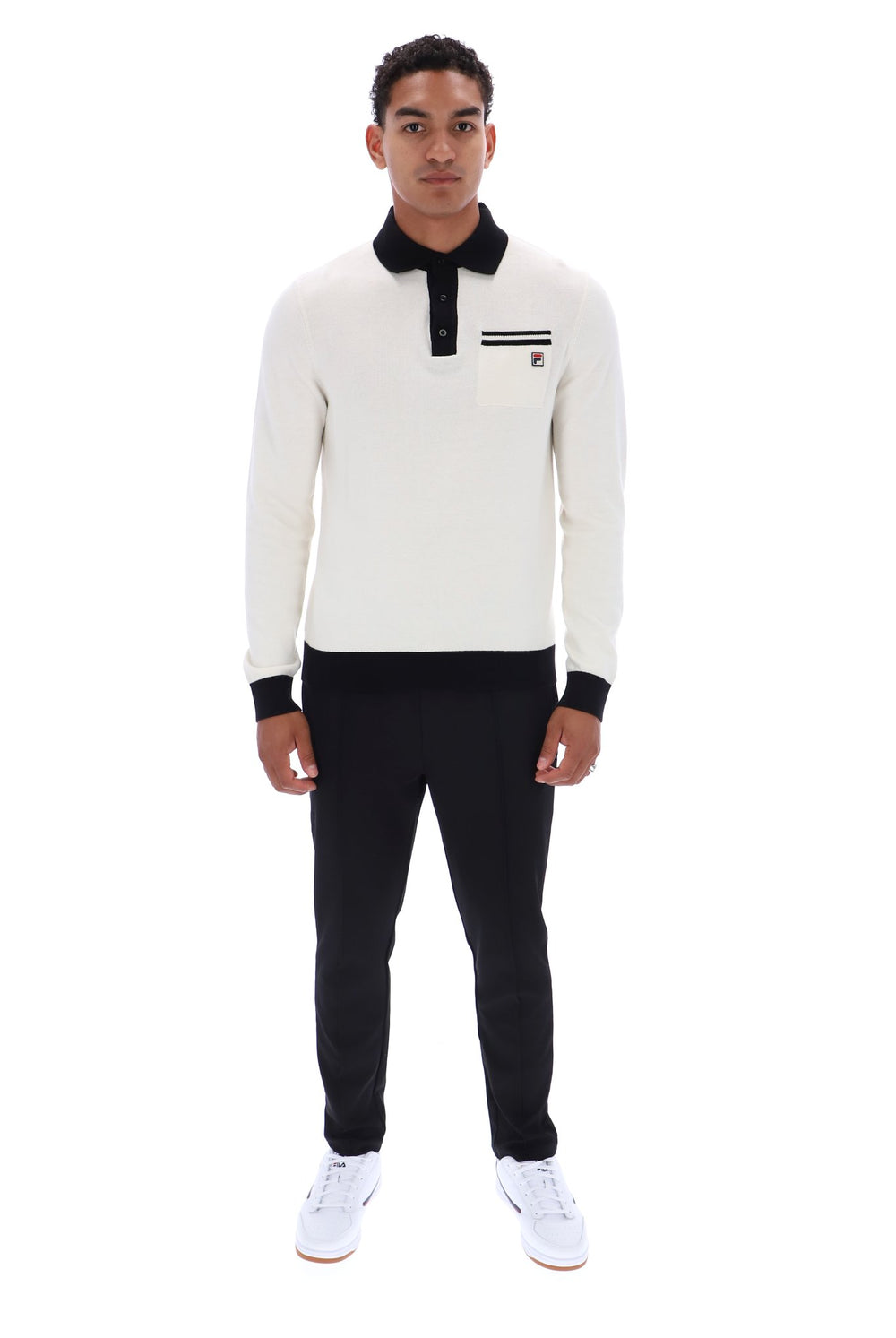 A man modeling a long-sleeve knit polo shirt in off-white with black collar, cuffs, and placket, paired with black trousers and white sneakers, shot against a plain white background.