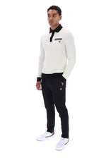 A man modeling a white long-sleeve polo shirt with a black collar and chest stripe detail, paired with black jogger pants and white sneakers, against a plain white background.
