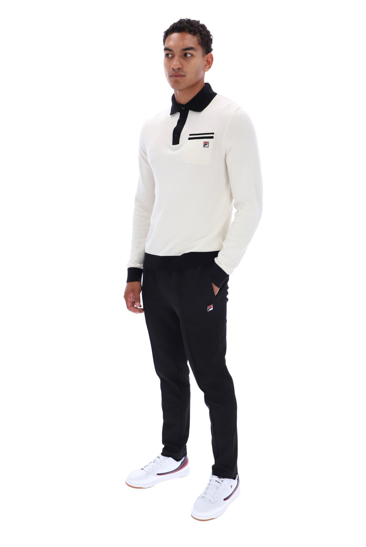 A man modeling a white long-sleeve polo shirt with a black collar and chest stripe detail, paired with black jogger pants and white sneakers, against a plain white background.