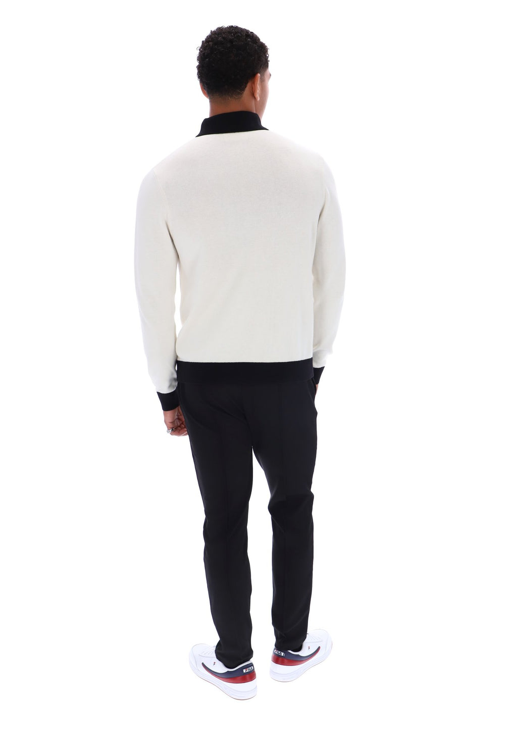 A man viewed from behind wearing a cream knit sweater with black ribbed trim, black trousers, and white sneakers with red accents, set against a plain white background.