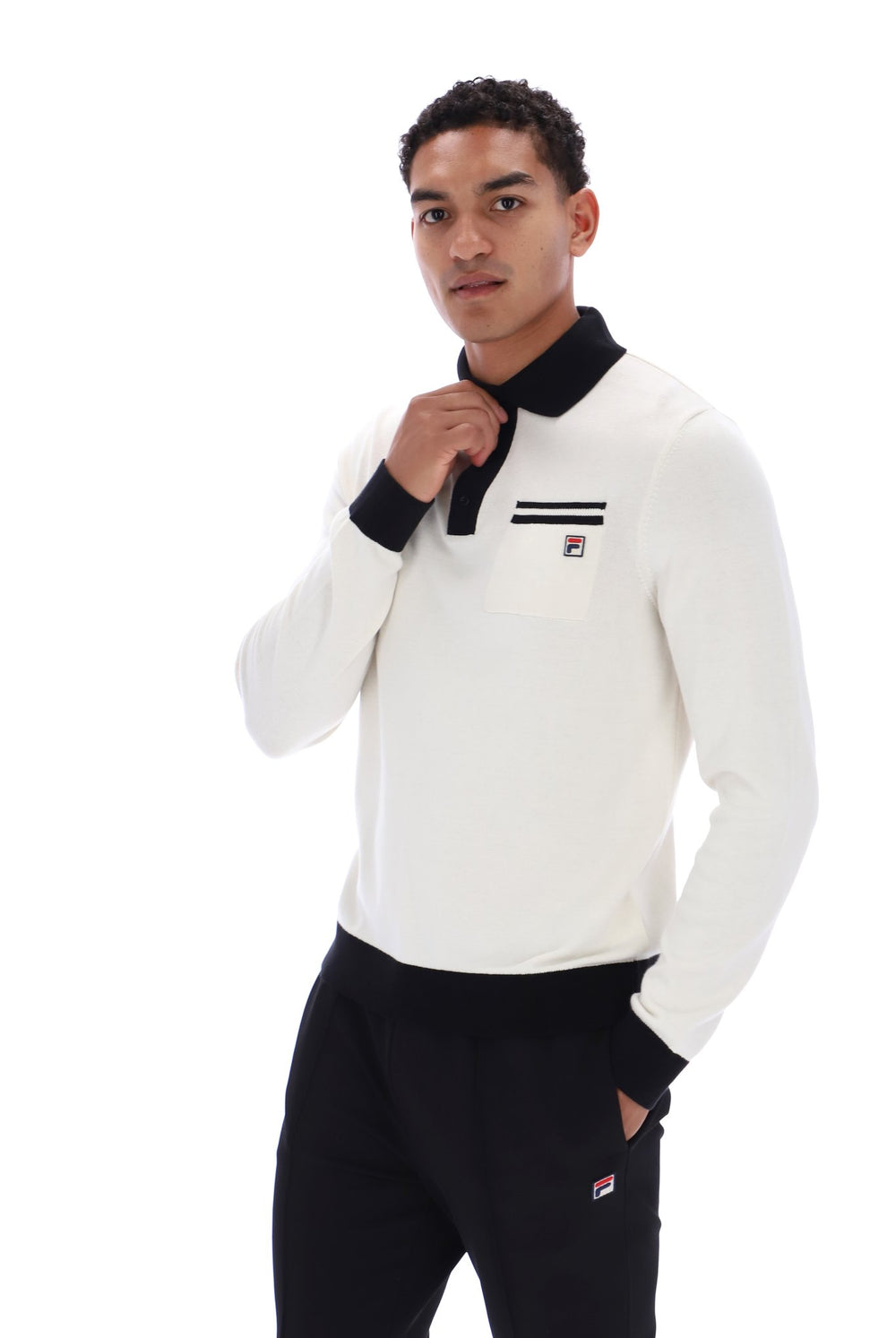 A man wearing a white long-sleeve knit polo shirt with a black collar and cuffs, featuring a small red, white, and blue Fila logo on the left chest, paired with black sweatpants against a white background.