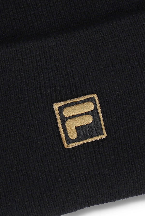 Close-up of a black ribbed knit beanie with a gold embroidered square FILA logo patch on the cuff.