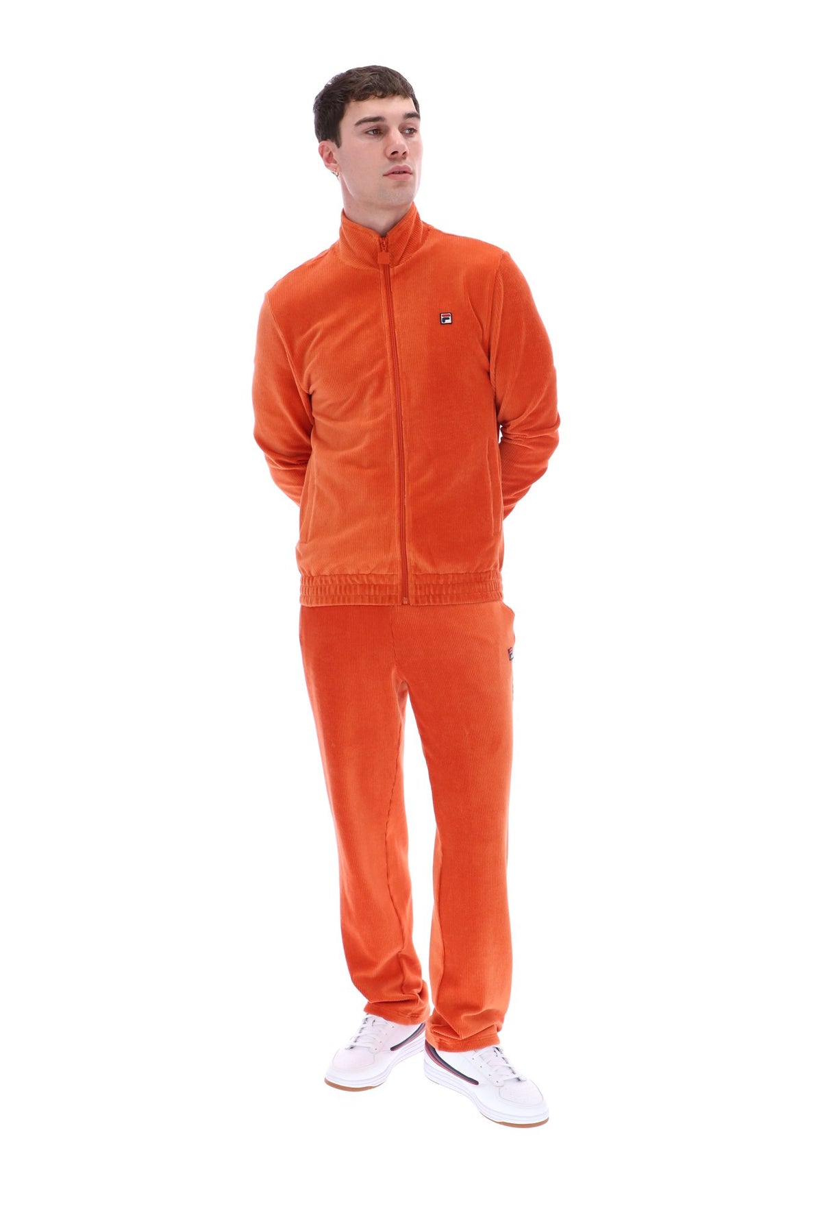 A man modeling a matching orange corduroy tracksuit, consisting of a zip-up jacket and matching pants, paired with white sneakers, against a plain white background.