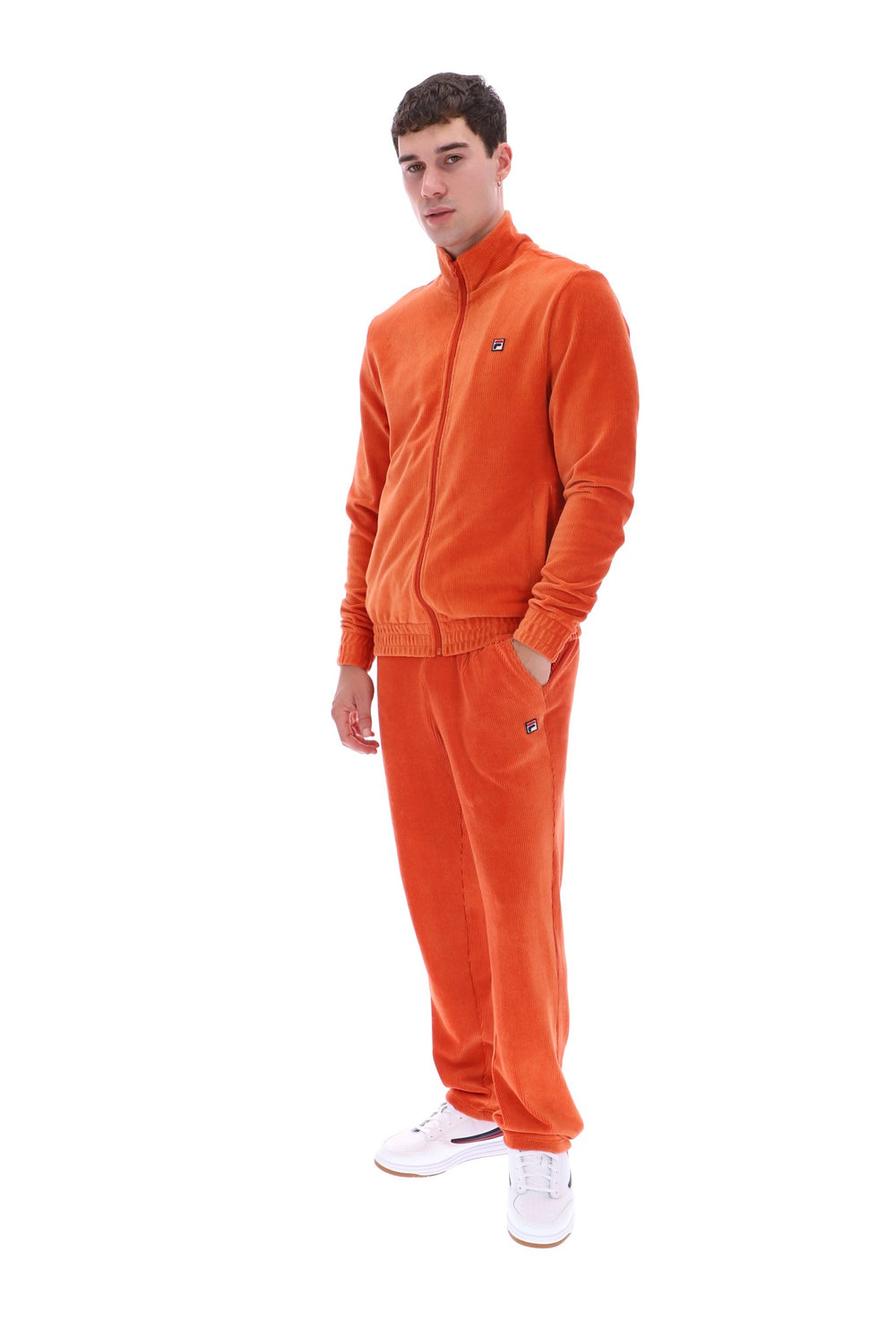 A man modeling a bright orange corduroy tracksuit, consisting of a zip-up jacket and matching sweatpants, paired with white sneakers, against a solid white background.