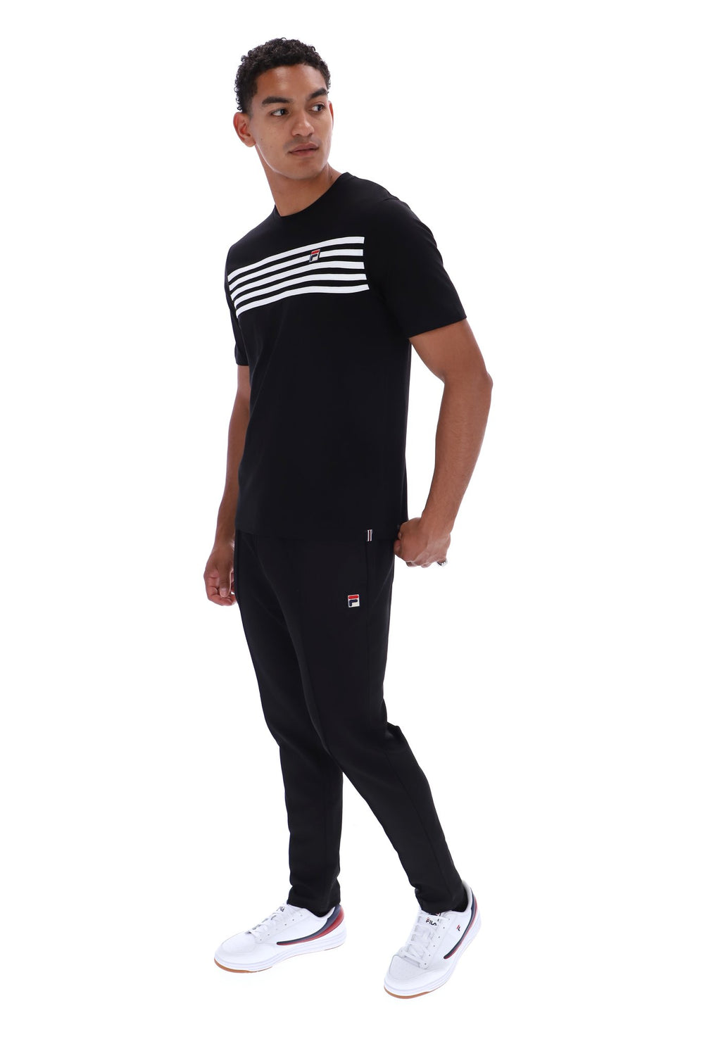 A man modeling a black short-sleeve t-shirt with three white horizontal stripes across the chest, paired with black joggers featuring a small red and white logo on the left thigh, and white sneakers, set against a plain white background.