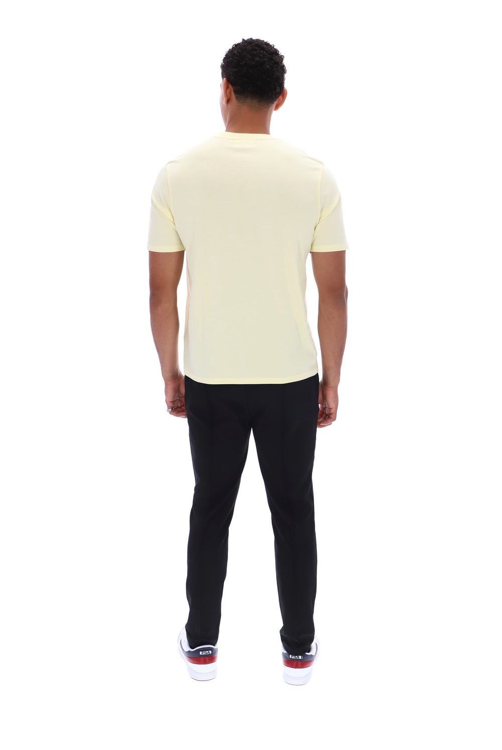 A man viewed from behind wearing a pale yellow short-sleeve t-shirt, black pants, and white sneakers with red accents, set against a plain white background.