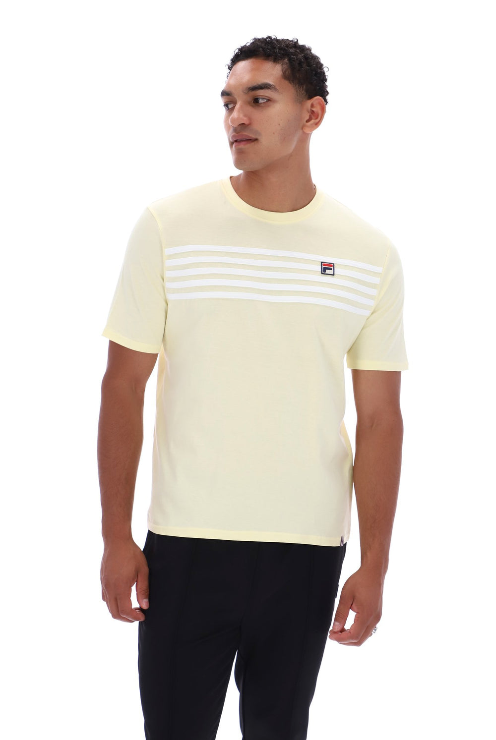 A man wearing a pale yellow short-sleeve t-shirt with white horizontal stripes across the chest and a small Fila logo on the left chest, paired with black pants, against a plain white background.