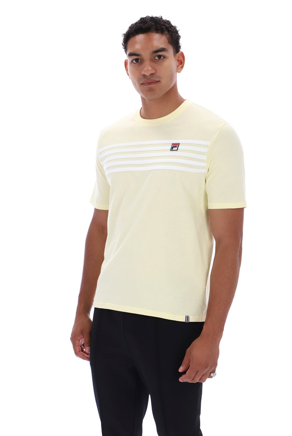 A man wearing a pale yellow Fila short-sleeve t-shirt with white horizontal stripes across the chest and a small red and blue logo, paired with black pants, against a plain white background.