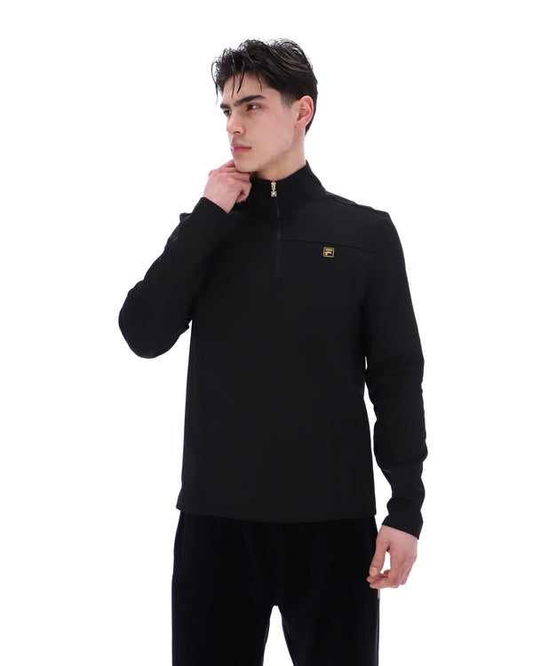 Man wearing a black jacket with a FILA gold logo, paired with matching bottoms, against a white background