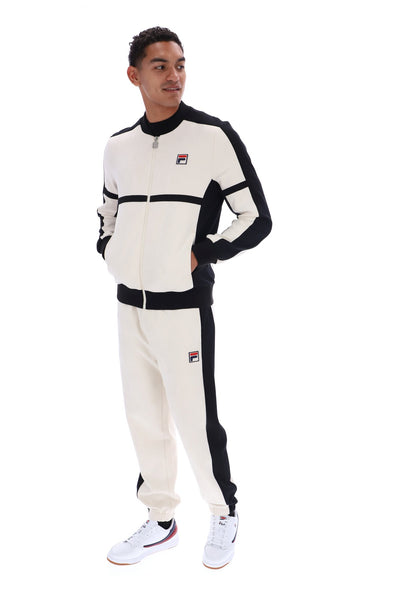 A man modeling a Fila tracksuit featuring a cream and black zip-up jacket and matching jogger pants, paired with white sneakers, against a solid white background.