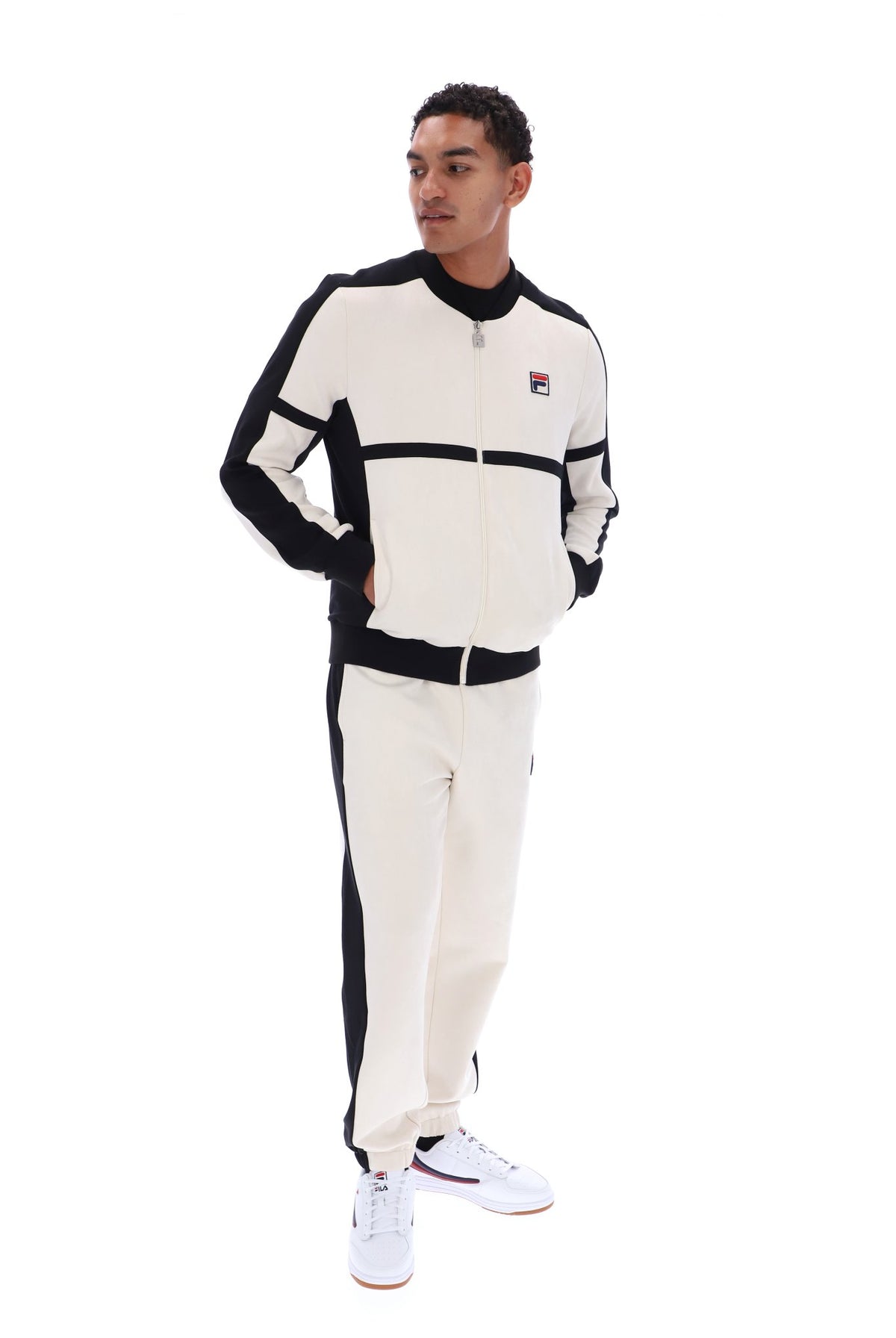 A man modeling a matching cream and black Fila tracksuit, featuring a zip-up jacket with a small Fila logo on the chest and matching jogger pants, set against a plain white background.