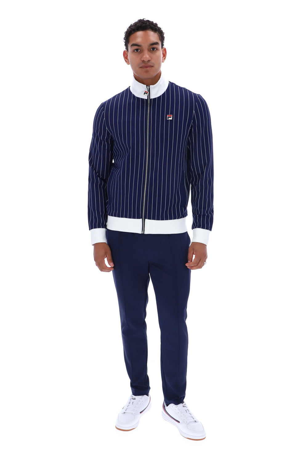 A man modeling a navy blue pinstriped zip-up jacket with white ribbed trim, matching navy pants, and white sneakers against a plain white background.