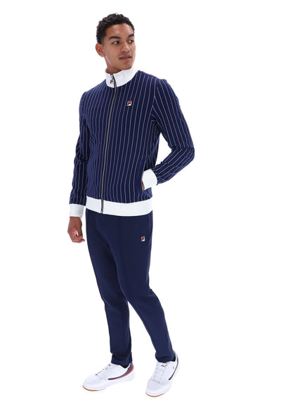 A man modeling a navy blue pinstripe tracksuit with white accents, including a zip-up jacket and matching sweatpants, paired with white sneakers, on a solid white background.