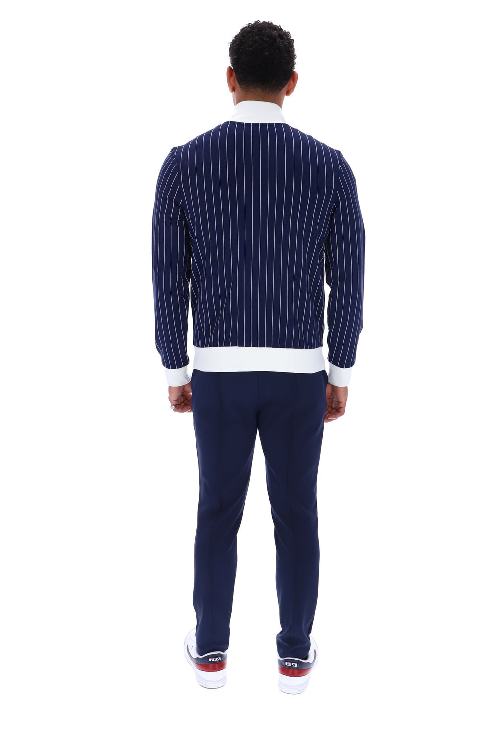 A man viewed from behind wearing a navy blue pinstriped sweater with white ribbed trim, navy pants, and white sneakers with red accents, set against a plain white background.